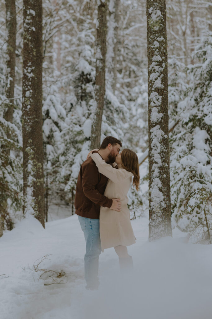 Couple embracing during a winter engagement session in a snow-covered pine forest in St. Germain, Wisconsin