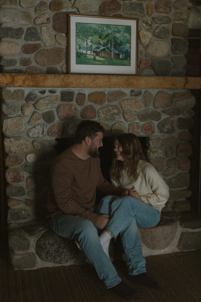 Couple sitting together by a stone fireplace inside a rustic cabin during a winter engagement session in Wisconsin