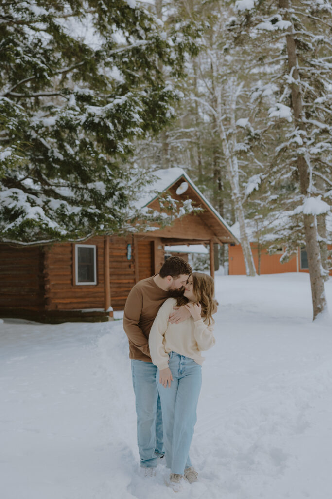 Couple sharing a kiss outside a snow-covered cabin during a winter engagement session in Northern Wisconsin