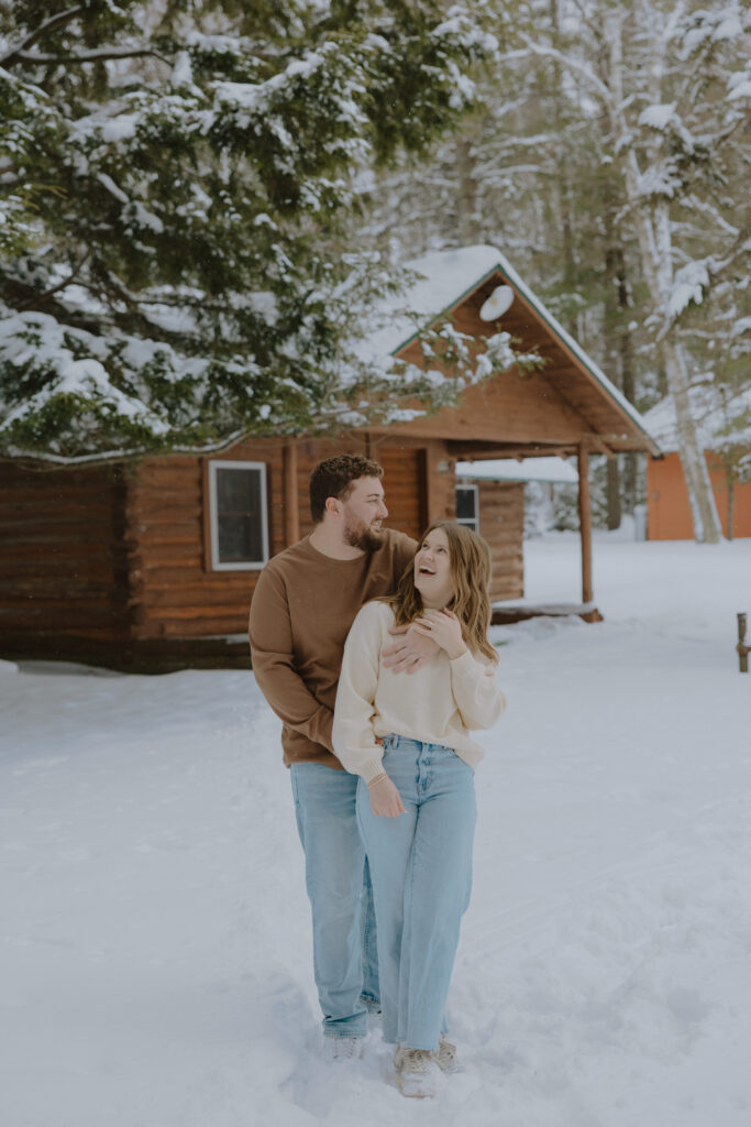 Couple laughing together outside a cozy cabin during a winter engagement session in Northern Wisconsin