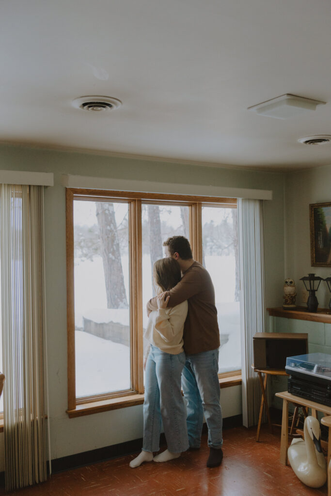 Couple embracing by a window inside a cabin during a winter engagement session in Northern Wisconsin